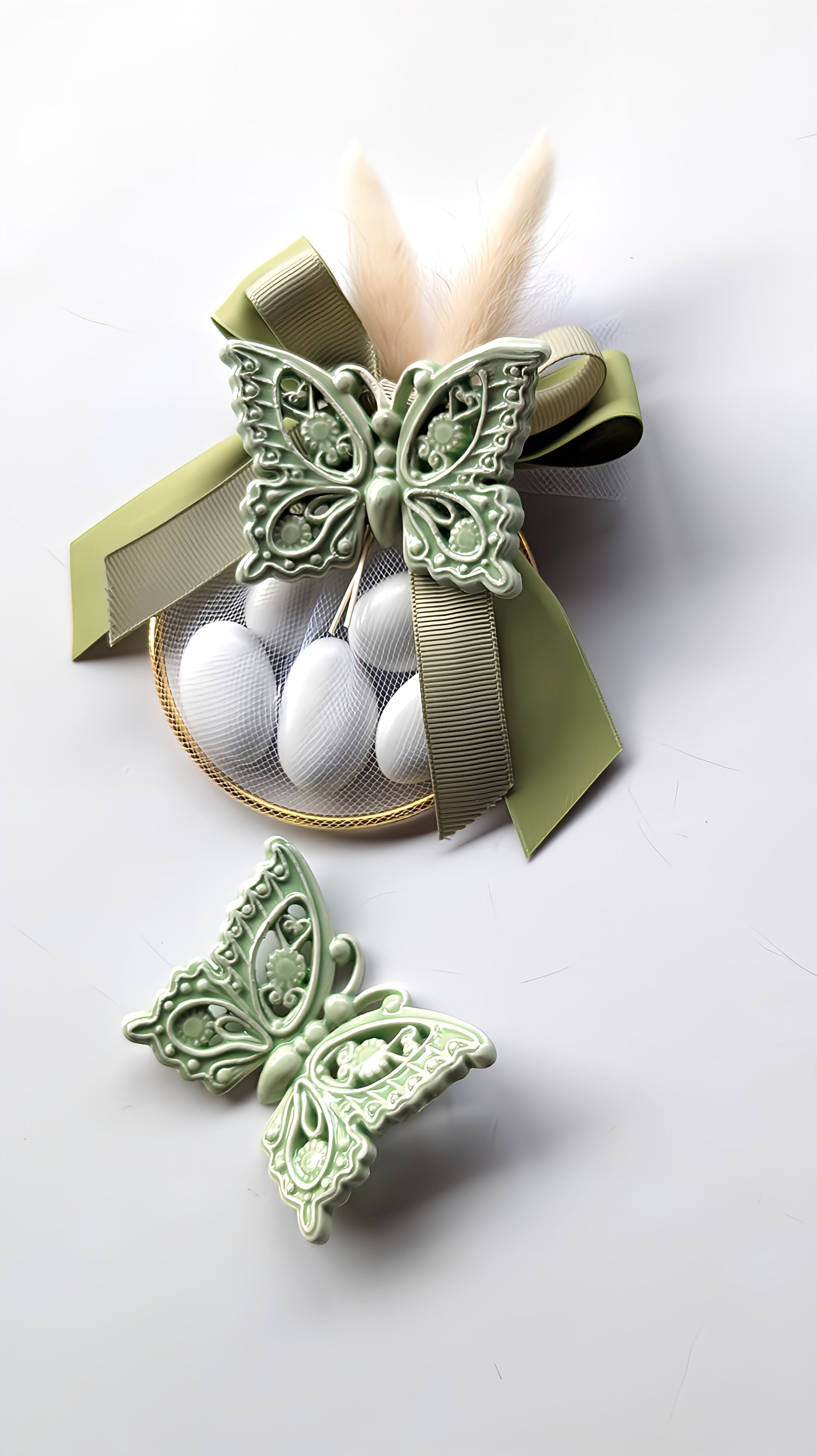 Bomboniera artigianale con farfalla in ceramica verde salvia, confezionata con tulle bianco, cinque confetti, cerchio dorato, fiocco doppio in raso e piuma decorativa su sfondo chiaro.