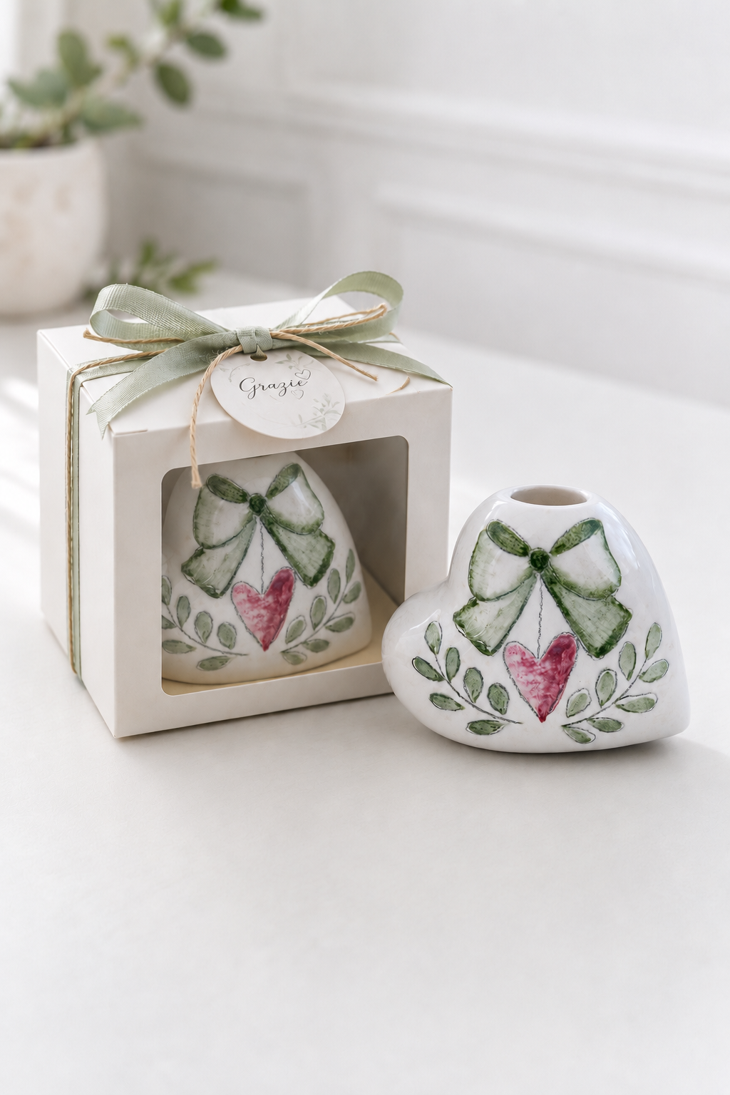 Profumatore a cuore in ceramica decorato a mano con fiocco verde salvia e cuore rosa, presentato accanto alla confezione regalo con finestra.